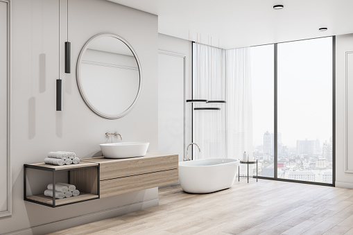 Stylish spacious bathroom in the morning with round mirror above sink, wooden tabletop and floor, white bath near huge window with city view. 3D rendering