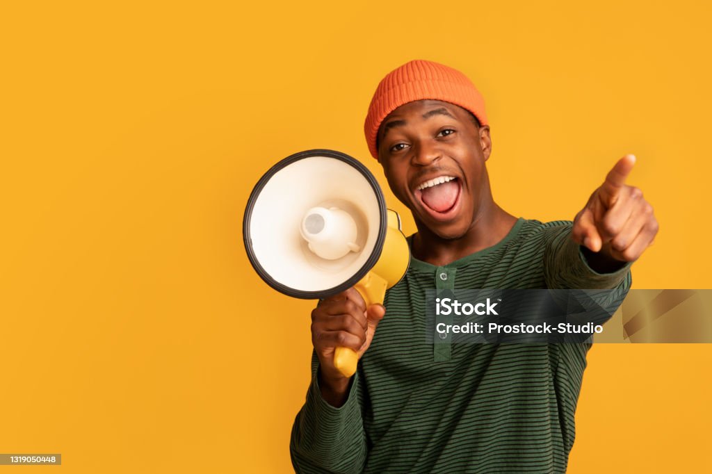 Cheerful Black Guy Shouting In Loudspeaker And Pointing At Camera Cheerful Black Guy Shouting In Loudspeaker And Pointing At Camera, Excited African American Man Holding Megaphone, Making Announcement, Standing Isolated Over Yellow Background, Copy Space Megaphone Stock Photo
