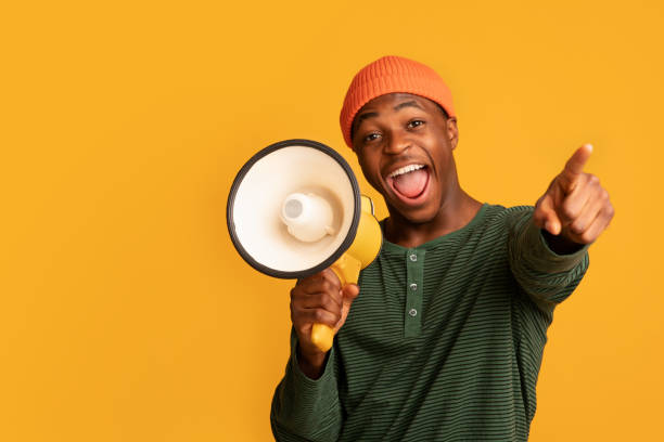 Cheerful Black Guy Shouting In Loudspeaker And Pointing At Camera Cheerful Black Guy Shouting In Loudspeaker And Pointing At Camera, Excited African American Man Holding Megaphone, Making Announcement, Standing Isolated Over Yellow Background, Copy Space black orange audio stock pictures, royalty-free photos & images