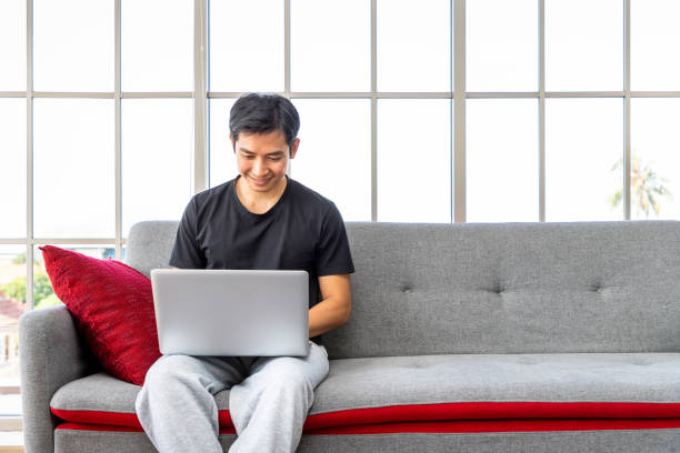 Happy young Asian man using laptop computer sitting o sofa in living room, work from home concept.