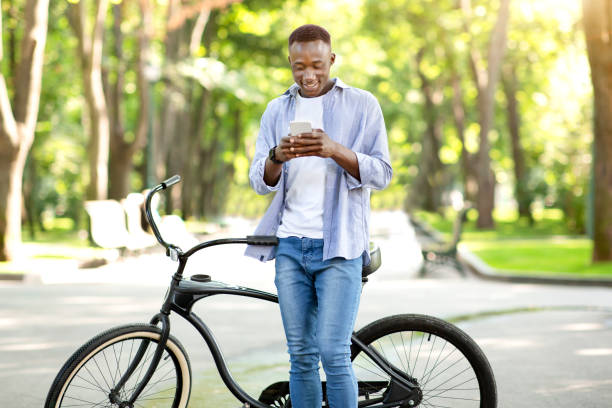 istock Funky black guy with bicycle sending communicating online on smartphone at park 1319050726