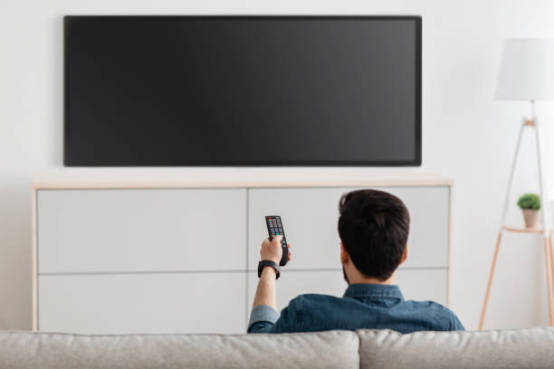istock Man watching television, turning on plasma flatscreen TV-set, pointing remote control at empty TV screen on white wall 1319050751