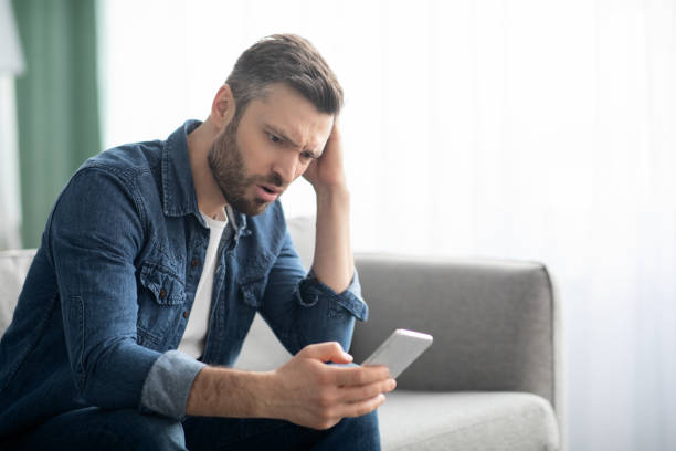 istock Closeup of thoughtful man sitting on couch, using smartphone 1319051532