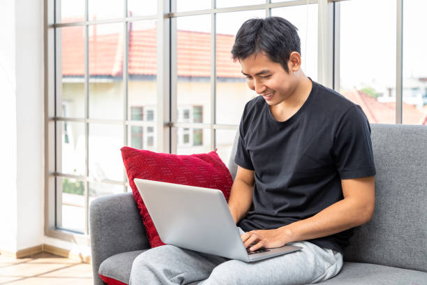 istock Happy young Asian man using laptop computer sitting o sofa in living room, work from home concept. 1319051625