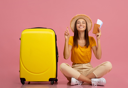 Happy young woman in causal outfit with passport and flight tickets sitting by yellow suitcase, closing eyes and crossing fingers, waiting for vacation, pink studio background, copy space