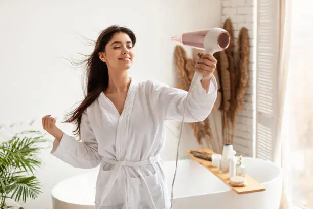 Haircare. Brunette Woman Drying And Styling Hair With Hairdryer Making Hairstyle Standing In Modern Bathroom At Home. Happy Female Using Hair Dryer Enjoying Beauty Routine After Morning Shower