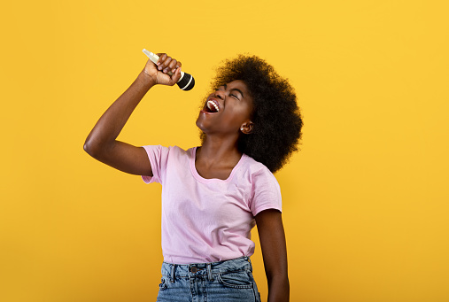 Joyful african american lady singing songs in microphone, having fun and enjoying the moment, having new hobby, standing over yellow background, studio shot. Lifestyle concept