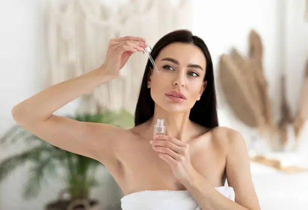 Modern beauty products at home, cosmetology, anti-aging cosmetics and advice from blogger and specialist. Young brunette lady in towel applies oil to skin of face, on bedroom interior background