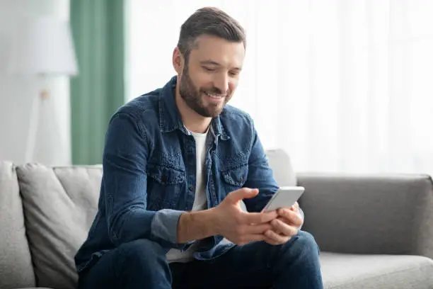 Cheerful middle-aged man sitting on sofa with modern mobile phone at home, chatting with friends, copy space. Positive bearded man using smartphone, checking new mobile applications