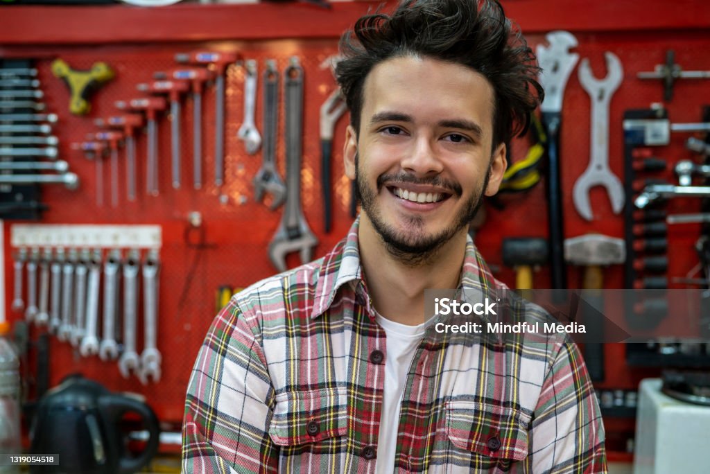 Smiling young repair mechanic standing with arms crossed in bicycle repair shop Portrait of smiling young man standing in front of tool rack indoors Bicycle Stock Photo