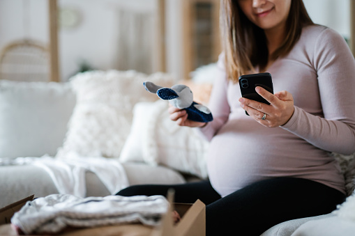 Cropped shot of Asian pregnant woman relaxing on sofa, shopping online with smartphone. She has unboxed a delivery package of baby clothing and toys and holding a bunny toy rattle. Time to get some baby essentials for the unborn baby!