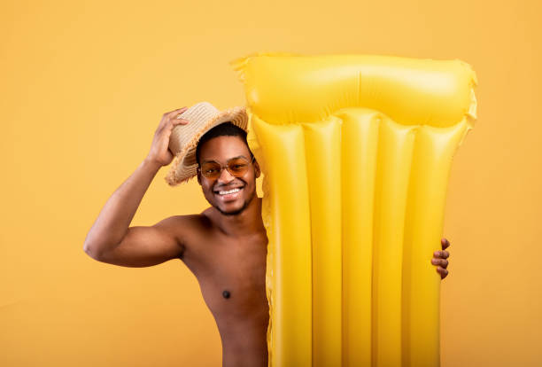 istock Hello summer. Black man lifting his straw hat in greeting gesture, hiding behind inflatable lilo on yellow background 1319057271