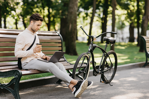 Work with pleasure. Side view of handsome man with stubble in casual clothes with bag, typing on laptop and drinking coffee takeaway, sitting on bench near bicycle in city park, outdoors, free space