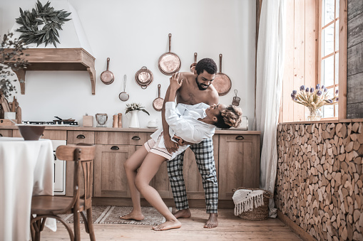 Life style. African american man shirtless and pretty woman in shirt barefoot dancing in morning at home in kitchen