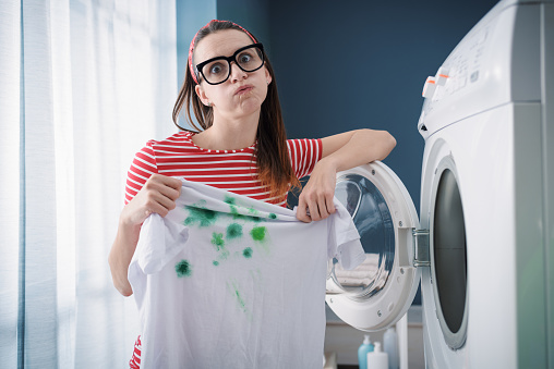 Young disappointed woman holding stained clothes and standing in front of the washing machine, laundry problems concept