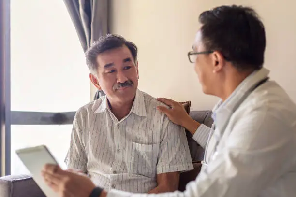 Smiling doctor consoling senior adult patient in medical clinic. Kind caring therapist or caregiver helping older retired man talking, giving comfort, expressing care concept.