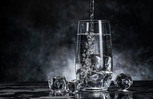 Pouring water from bottle into glass with ice cubes on black background