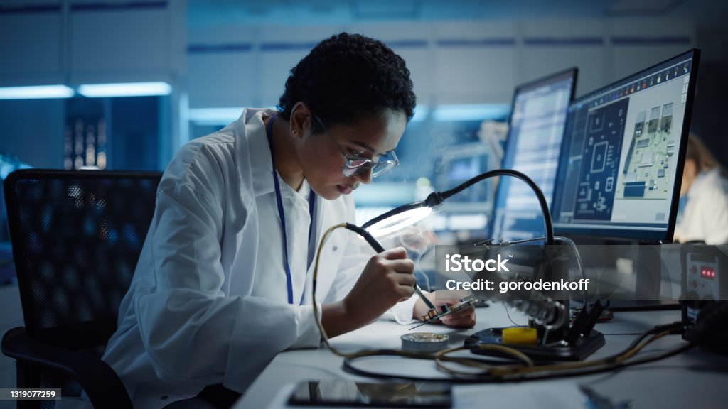Modern Electronics Research, Development Facility: Black Female Engineer Does Computer Motherboard Soldering. Scientists Design PCB, Silicon Microchips, Semiconductors. Medium Close-up Shot Engineer Stock Photo