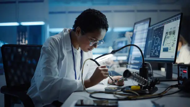 Photo of Modern Electronics Research, Development Facility: Black Female Engineer Does Computer Motherboard Soldering. Scientists Design PCB, Silicon Microchips, Semiconductors. Medium Close-up Shot
