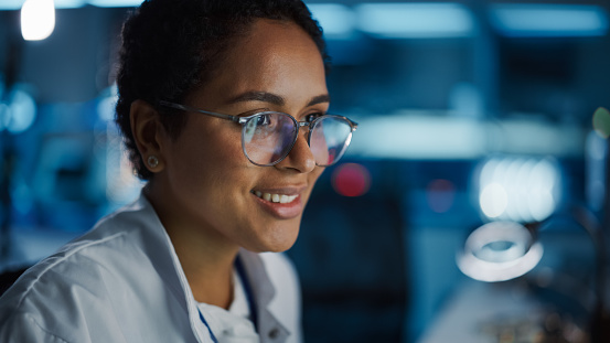 Portrait of Beautiful Black Latin Woman Computer Screen Reflecting in Her Glasses. Young Intelligent Female Scientist Working in Laboratory. Background Bokeh Blue with High-Tech Technological Lights