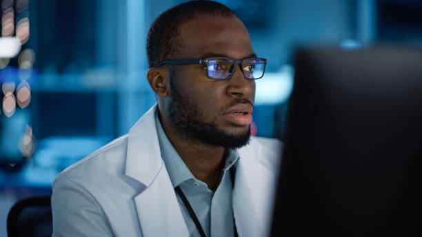 ritratto di bell'uomo nero che indossa occhiali che lavorano con sicurezza su un computer. giovane ingegnere o scienziato intelligente che lavora in laboratorio. sfondo blu bokeh. colpo ravvicinato - young adult engineer intelligence male beauty foto e immagini stock