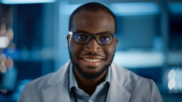 istock Handsome Black Man Wearing Glasses Smiling Charmingly Looking at Camera. Young Intelligent Male Engineer or Scientist Working in Laboratory. Technological Bokeh Blue Background. Close-up Shot 1319077833