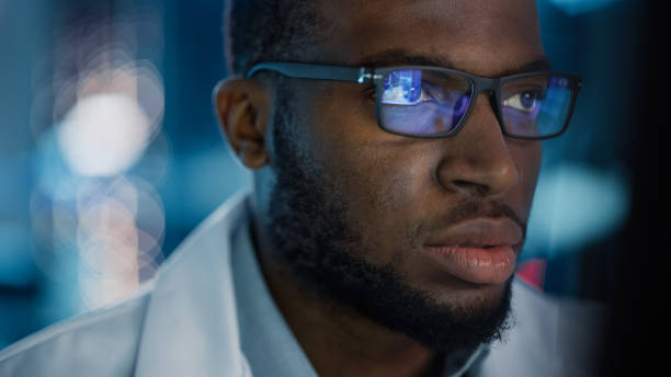 ritratto di bell'uomo nero che indossa occhiali che lavorano con sicurezza su un computer. giovane ingegnere o scienziato intelligente che lavora in laboratorio. sfondo blu bokeh. colpo ravvicinato - young adult engineer intelligence male beauty foto e immagini stock