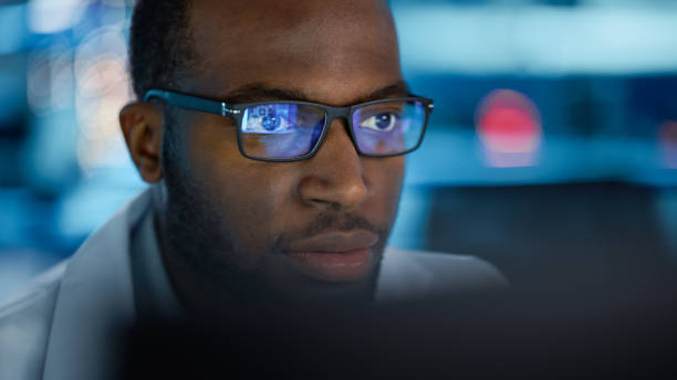 istock Portrait of Handsome Black Man Wearing Glasses Working Confidently on a Computer. Young Intelligent Male Engineer or Scientist Working in Laboratory. Bokeh Blue Background. Close-up Shot 1319077924