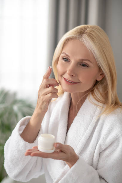 istock A woman in a white bath robe applying facial cream on her cheeks 1319085049