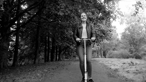 Black and white portrait of smiling young woman enjoys riding on kick scooter at park.