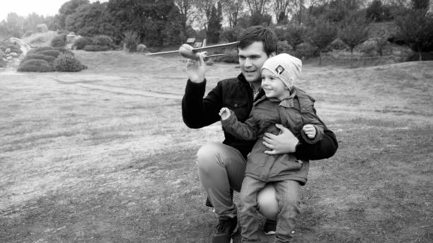 retrato preto e branco de jovem pai com filho pequeno jogando avião de brinquedo no campo no parque - airplane black and white fun child - fotografias e filmes do acervo
