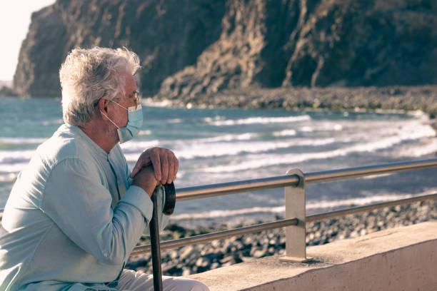 istock Coronavirus. Bearded old man sitting on the beach in the sunset light holding a walking stick because of body aches 1319087062
