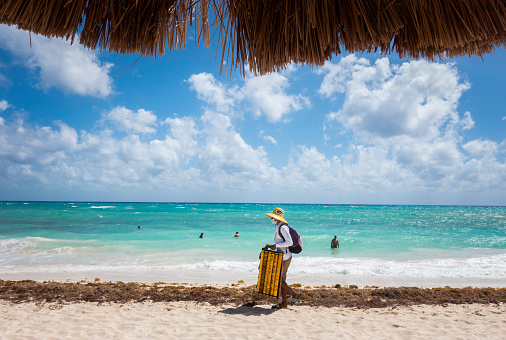 Playa del Carmen, Mexico - March 1, 2021: A Hispanic female wearing a face mask and straw hat walks along the beach selling handmade bracelets to tourists vacationing during the Covid pandemic.
