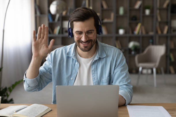istock Smiling man in headphones waving hand at webcam, using laptop 1319095644