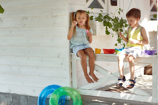 caucasian boy and girl sitting on a porch playing with paper boats, making soap bubbles