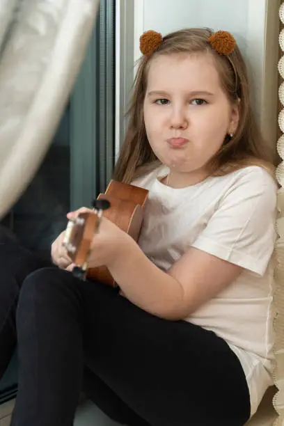 Photo of Portrait of sad upset little girl in white t-shirt and black leggings sitting inside of home and playing ukulele.