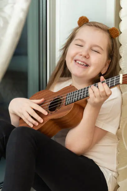 Photo of Portrait of happy smiling little girl in white t-shirt and black leggings sitting inside of home and playing ukulele.