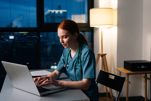 felice sorridente giovane medico donna in uniforme medica verde blu che lavora digitando sul computer portatile. - lavoro straordinario foto e immagini stock