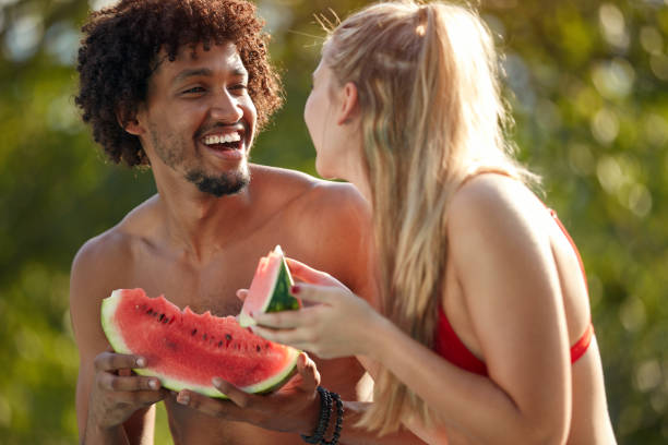 multiethnic couple looking each other, smiling, laughing, while eating watermelon young multiethnic couple looking each other, smiling, laughing, while eating watermelon black man eating watermelon stock pictures, royalty-free photos & images