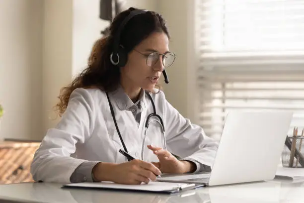 Photo of Focused young female doctor in headset watching webinar taking notes