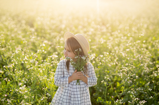 A beautiful girl in a hat and with a bouquet of flowers in the sun gently holds a bouquet of flowers. The child looks away with tenderness and love. Peace, tranquility and unity with nature