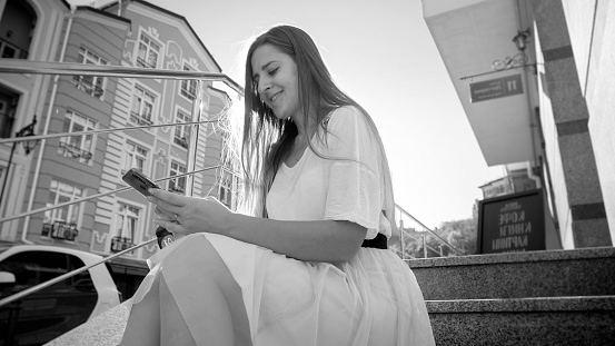 Black and white portrait of smiling female student with smartphone sitting on stone stairs on the city street.