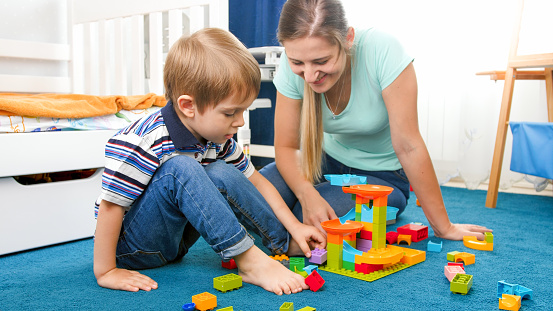 Beautiful young woman helping her toddler son building colorful plastic constructor.