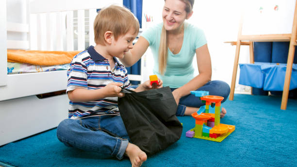 nette lächelnde kleinkind junge aufräumen sein zimmer und holen spielzeug in der tasche - child playroom parent indoors stock-fotos und bilder