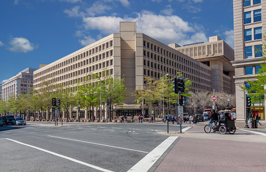 Federal Bureau of Investigation (FBI) Headquarters on Pennsylvania Avenue, Washington DC, USA. Blue Sky with Puffy clouds, Street, Rickshaw, Passersby and Green Trees are in the image. Wide angle lens.