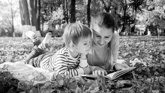 Black and white portrait of family reading story book while lying on ground at park.