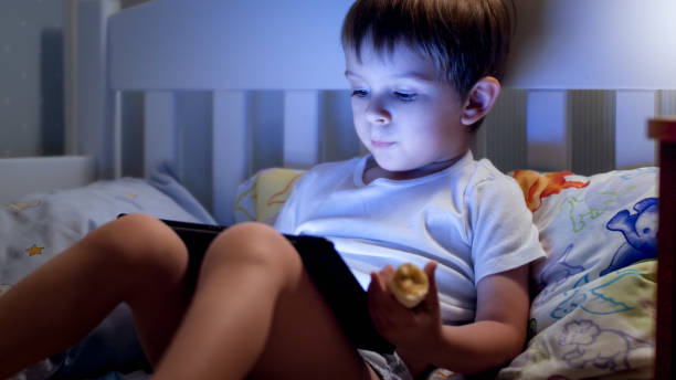 Portrait of cute little boy lying in bed at night and using digital tablet computer