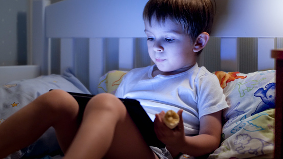 Portrait of cute little boy lying in bed at night and using digital tablet computer.
