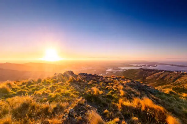 This May 2021 image shows an autumn sunset over Ōtautahi Christchurch, Aotearoa New Zealand on a clear day. The city is the largest on Te Waipounamu South Island. Photographed from near the Gondola in Mt. Pleasant in the Port Hills along the Banks Peninsula. The Avon Heathcote Estuary is seen on the right where the Avon River meets the Pacific Ocean. A thin layer of fog hangs in the distance.