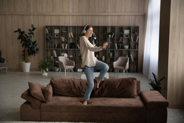 istock Overjoyed female student jump on sofa by music in headset 1319136483